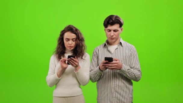 Um homem e uma mulher estão de pé e a enviar mensagens num smartphone. Uma mulher está a olhar para o telemóvel dela. Eles estão de pé sobre um fundo verde. Tela verde. 4K — Vídeo de Stock