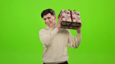 Adam hediye kutusunu kulağının yanında sallıyor. - İçinde ne olduğunu bilmek istiyor. Gülümsüyor. O, yeşil bir arka planın üzerinde duruyor. Yeşil ekran. 4K