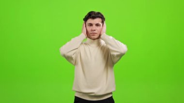 Adam ayakta duruyor ve kameraya bakıyor. Kulaklarını, ağzını ve gözlerini elleriyle kapatıyor. O, yeşil bir arka planın üzerinde duruyor. Yeşil ekran. 4K