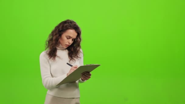 Η γυναίκα στέκεται και κοιτάζει την κάμερα. Κρατάει σημειώσεις στο έγγραφο. Είναι συγκεντρωμένη. Στέκεται πάνω σε ένα πράσινο φόντο. Πράσινη οθόνη. 4K — Αρχείο Βίντεο