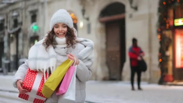 Seorang wanita pergi ke pusat kota saat salju turun. Dia memegang hadiah besar dan tas belanja. Dia tersenyum dan melihat ke kamera. Pengambilan gambar. 4K — Stok Video