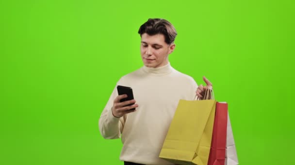Um homem está enviando mensagens em um smartphone e segurando sacos de compras em suas mãos. Ele está levantando a cabeça e sorrindo. Ele está de pé sobre um fundo verde. Tela verde. 4K — Vídeo de Stock