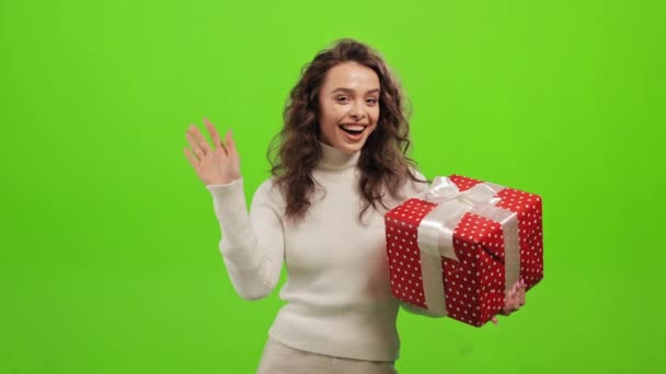 Женщина стоит и смотрит в камеру. Она держит подарок и машет рукой. Она стоит на зеленом фоне. Зеленый экран. 4K — стоковое видео