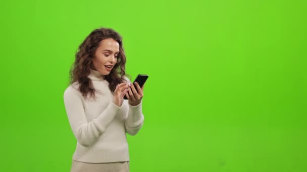 Η γυναίκα ψάχνει μέσω των κοινωνικών δικτύων στο smartphone της. Χαμογελάει και κοιτάζει την κάμερα. Στέκεται πάνω σε ένα πράσινο φόντο. Πράσινη οθόνη. 4K — Αρχείο Βίντεο