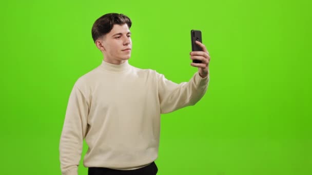 Mężczyzna robi sobie selfie na smartfonie. Wkłada rękę do kieszeni. Stoi na zielonym tle. Zielony ekran. 4K — Wideo stockowe