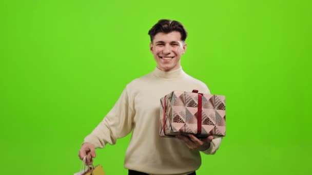 The man is looking at the camera and smiling. He is holding a gift and picking up shopping bags. He is standing on a green background. Green screen. 4K — Stock Video