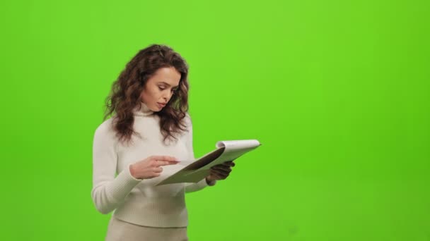 Kadın evrakları karıştırıyor ve gerekli bilgileri arıyor. Başını kaldırıyor ve kameraya bakıyor. O, yeşil bir arka planın üzerinde duruyor. Yeşil ekran. 4K — Stok video