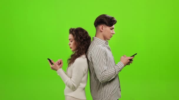 Un hombre y una mujer están de pie de espaldas el uno al otro y mensajes de texto en un teléfono inteligente. Una mujer está mirando en su teléfono inteligente mans. Están parados sobre un fondo verde. Pantalla verde. 4K — Vídeos de Stock