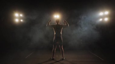 A man is dressing in tights is standing on the stage. He is waving his hands. The spotlight is shining on him. Shooting from behind. 4K