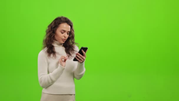 La mujer está mirando a través de las redes sociales en su teléfono inteligente. Está de pie sobre un fondo verde. Pantalla verde. 4K — Vídeo de stock