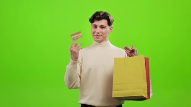 这个人看着相机笑着。他拿着购物袋，挥舞着信用卡。他站在绿色的背景上.绿色屏幕。4K — 图库视频影像