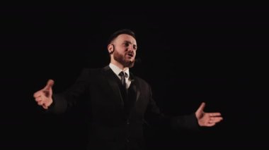 Adam sahnede duruyor ve duygusal olarak konuşuyor. O, ellerini sallıyor. Üzerinde bir mikrofon var. Takım elbise giyiyor. 4K