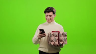 Bir adam akıllı telefondan mesaj atıyor ve elinde bir hediye tutuyor. Gülümsüyor ve el sallıyor. O, yeşil bir arka planın üzerinde duruyor. Yeşil ekran. 4K