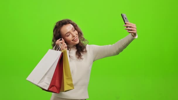 한 여성 이 스마트폰으로 셀카를 들고 쇼핑백을 손에 들고 있습니다. 웃고 있어요. 그녀는 녹색 배경 위에 서 있습니다. 녹색 화면. 4K — 비디오