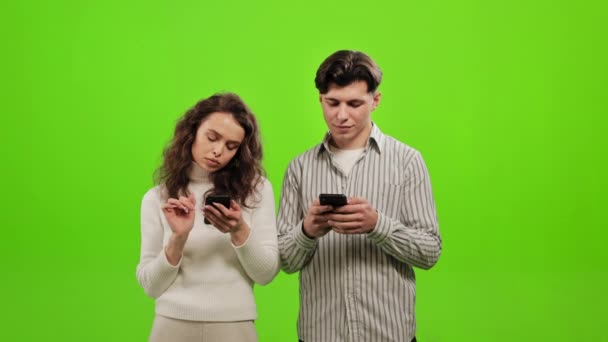 Um homem e uma mulher estão de pé e a enviar mensagens num smartphone. Eles estão de pé sobre um fundo verde. Tela verde. 4K — Vídeo de Stock