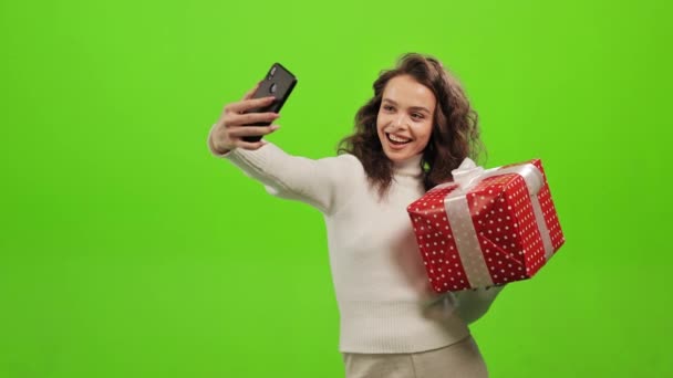 Kvinnan står och tittar på kameran. Hon håller i en gåva och tar en selfie på sin smartphone. Hon står på en grön bakgrund. Grön skärm. 4K — Stockvideo