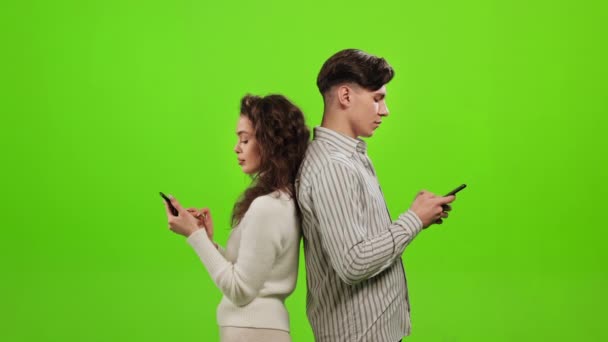 A man and a woman are standing with their backs to each other and texting on a smartphone. They are raising their heads and thinking. They are standing on a green background. Green screen. 4K — Stock Video