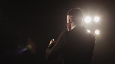 Konferansı konuşmacı yönetiyor. O, sahnede duruyor ve mikrofona konuşuyor. O, ellerini sallıyor. Spot ışıkları onun üzerinde parlıyor. Kamera onun etrafında dönüyordu. 4K