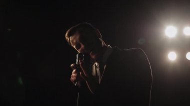 Şarkıcı şarkıyı duygusal olarak söylüyor. Elinde retro mikrofon var. Spot ışıkları onun üzerinde parlıyor. 4K