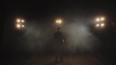 Bir adam sahnede duruyor ve seyirciye bakıyor. Kadehi onun üzerine kaldırır. Spot ışıkları onun üzerinde parlıyor. Kamera arkadan vurup kaçma olayı yapıyor. 4K