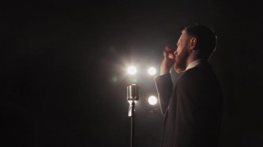 Bir erkek sahnede duygusal bir performans sergiliyor. Önünde bir mikrofon var. El sallıyor. Spot ışıkları onun üzerinde parlıyor. 4K
