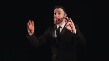 Konuşmacı sahnede duruyor ve duygusal olarak konuşuyor. O, ellerini sallıyor. Üzerinde bir mikrofon var. Spot ışıkları onun üzerinde parlıyor. Takım elbise giyiyor. 4K