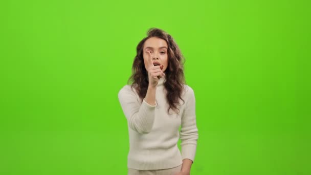 The woman is standing and looking at the camera. She is raising a finger to her lips and showing a gesture - quiet. She is standing on a green background. Green screen. 4K — Stock Video