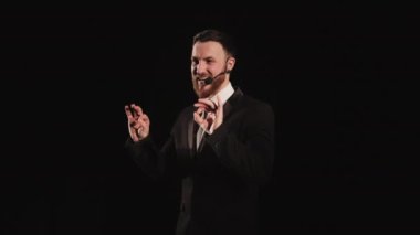 Konuşmacı sahnede duruyor ve konuşuyor. Üzerinde bir mikrofon var. Spot ışıkları onun üzerinde parlıyor. Takım elbise giyiyor. 4K