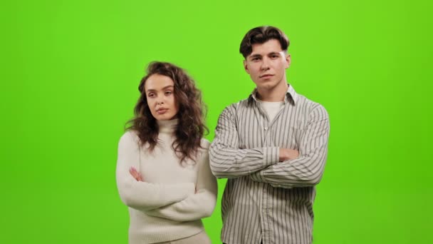 Un hombre y una mujer están de pie mirando a la cámara. Son serios y tienen los brazos cruzados. El hombre gira la cabeza y mira a la mujer. Están parados sobre un fondo verde — Vídeos de Stock