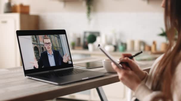 En kvinde sidder på sit hjemmekontor. Hun skriver notater i sin notesbog. Der er en bærbar computer på bordet. Hun taler på video med en mand. 4K – Stock-video