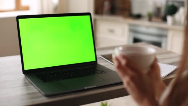 En kvinna sitter på sitt hemkontor. Hon håller i en kopp. På bordet finns en bärbar dator med en grön skärm. Det är Chromakey. 4K — Stockvideo