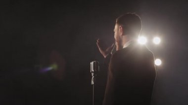 Sahnede bir adam sahne alıyor. Önünde bir mikrofon var. El sallıyor. Spot ışıkları onun üzerinde parlıyor. 4K