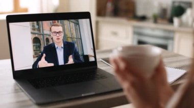 Bir kadın ev ofisinde oturuyor. O bir fincan tutuyor. Masada bir dizüstü bilgisayar var. Bir adamla videoda konuşuyor. 4K