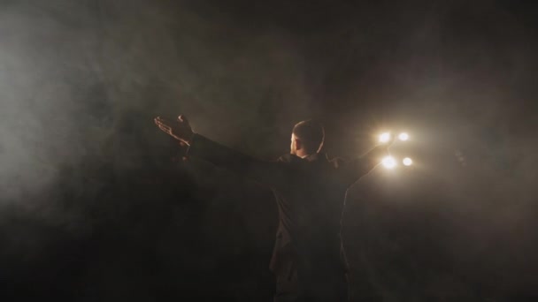 The speaker is standing on stage and holding up his hands. The spotlight is shining on him. Theres smoke all around it. 4K — Stock Video