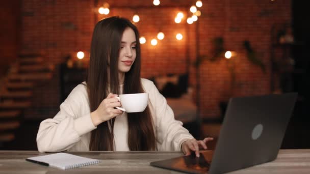 En kvinna sitter på sitt hemkontor. Hon sitter vid sitt skrivbord och jobbar på sin laptop. Hon dricker te. 4K — Stockvideo