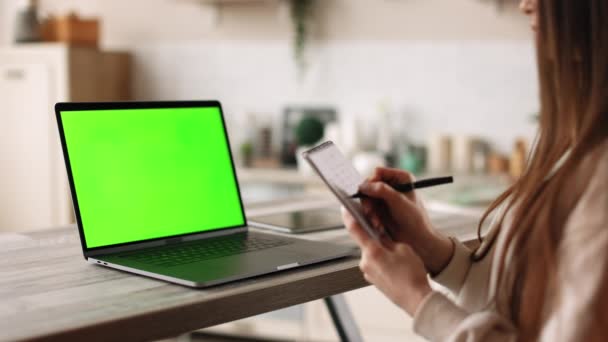 Μια γυναίκα κάθεται στο γραφείο του σπιτιού της. Κρατάει σημειώσεις στο σημειωματάριό της. Στο τραπέζι υπάρχει ένας φορητός υπολογιστής με πράσινη οθόνη. Χρωμάικι. 4K — Αρχείο Βίντεο