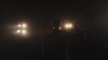 Adam sahnede duruyor ve mikrofonu kaldırıyor. Takım elbise giyiyor. Spot ışıkları onun üzerinde parlıyor. Etrafında duman var. 4K