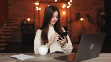 Bir kadın ev ofisinde oturuyor. O, masasında oturuyor ve akıllı telefonu üzerinde çalışıyor. O gülümsüyor. 4K