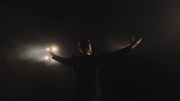 The speaker is standing on stage and holding up his hands. The spotlight is shining on him. Shooting from behind. 4K — Stock Video