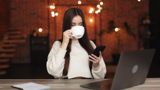 En kvinna sitter på sitt hemkontor. Hon dricker te och jobbar på sin smartphone. Det finns en laptop på bordet. 4K — Stockvideo
