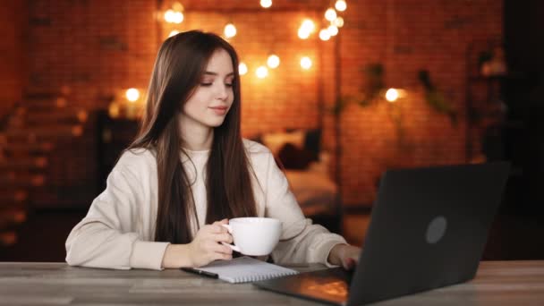 Uma mulher está sentada no seu escritório. Ela está trabalhando em um laptop e segurando uma xícara de chá. Ela está sorrindo. 4K — Vídeo de Stock