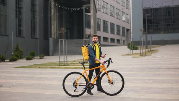 O entregador de comida está de pé e segurando o smartphone. Ele está a olhar para a câmara. Ele está segurando uma bicicleta e carregando uma grande mochila em seus ombros. Tiro de retrato. 4K — Vídeo de Stock