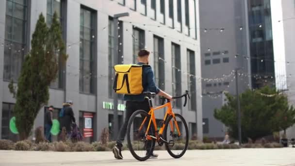 O tipo da entrega de comida vai. Ele está segurando uma bicicleta e carregando uma grande mochila em seus ombros. Por trás da vista. A câmara está a dar a volta. Tiro de retrato. 4K — Vídeo de Stock