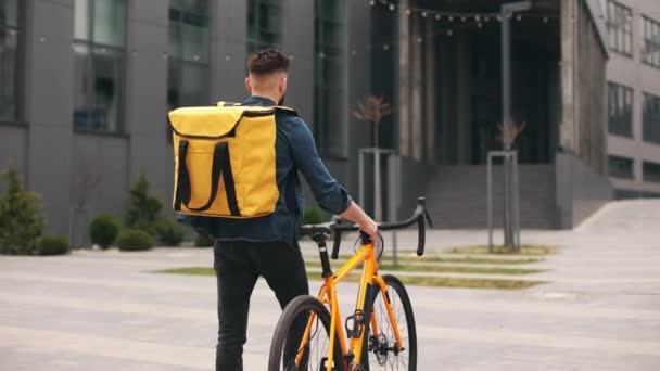 Der Essenslieferant geht und schaut auf das Smartphone. Er trägt ein Fahrrad und einen großen Rucksack auf den Schultern. Die Kamera schießt von hinten. Porträtschießen. 4K — Stockvideo