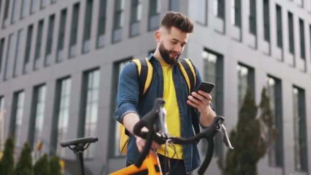 De bezorger staat naar de smartphone te kijken. Hij houdt een fiets vast en draagt een grote rugzak op zijn schouders. De camera is van onderen aan het fotograferen. Portret schieten. 4K — Stockvideo
