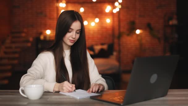 Bir kadın ev ofisinde oturuyor. O bir dizüstü bilgisayar üzerinde çalışıyor ve bir deftere notlar alıyor. 4K — Stok video