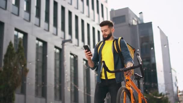 De bezorger gaat naar de smartphone kijken. Hij houdt een fiets vast en draagt een grote rugzak op zijn schouders. De camera is van onderen aan het fotograferen. Portret schieten. 4K — Stockvideo