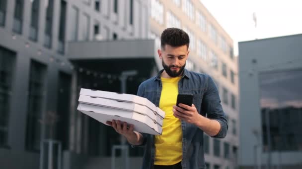 De bezorger staat te sms 'en op een smartphone. Hij houdt dozen pizza vast. Mooi Business Center op de achtergrond. Portretfoto 's. De camera beweegt. 4K — Stockvideo