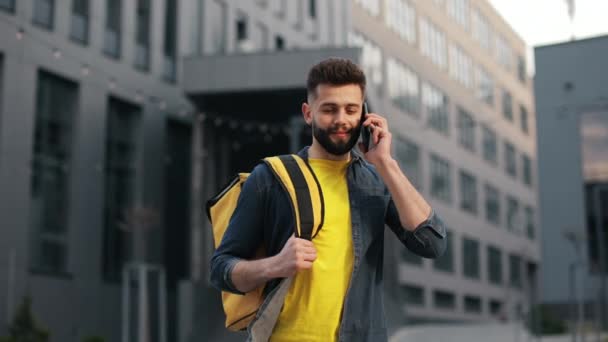 Der Essenslieferant steht und spricht auf einem Smartphone. Er trägt einen großen Rucksack auf den Schultern. Schöne Business Center im Hintergrund. Porträtaufnahmen. 4K — Stockvideo