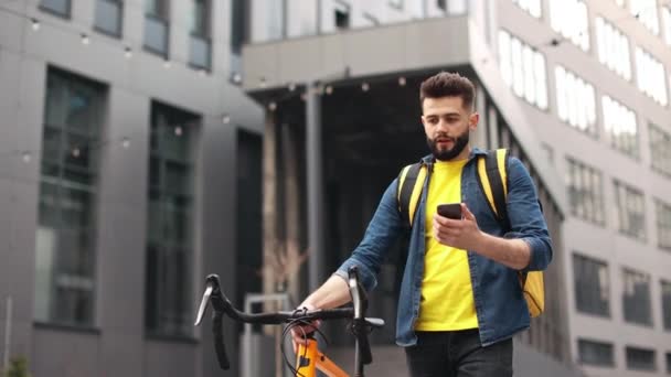 De bezorger gaat. Hij is serieus en kijkt naar de smartphone. Hij houdt een fiets vast en draagt een grote rugzak op zijn schouders. Mooi Business Center op de achtergrond. 4K — Stockvideo